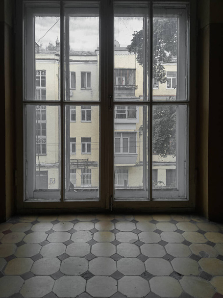 Θέα μέσα από ένα μεγάλο παράθυρο μιας σκοτεινής ερειπωμένης σκάλας ενός παλιού σπιτιού της σοβιετικής περιόδου του 1930 σε μια συννεφιασμένη μέρα. - Φωτογραφία, εικόνα