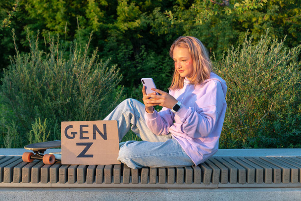 Ein stylisches Teenager-Mädchen in fliederfarbener Kapuzenjeans mit rosa Haaren sitzt auf einer Bank, das Telefon in der Hand, neben einem Longboard und einem Schild mit der Aufschrift Generation Z. GEN Z STYLE TRENDS. - Foto, Bild
