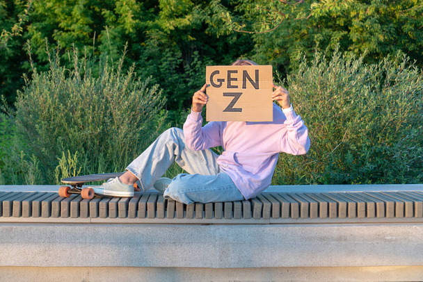 Elegante ragazza adolescente in jeans con cappuccio viola con capelli rosa si siede su una panchina accanto a un longboard il suo viso è coperto con un poster con l'iscrizione Generation Z. GEN Z Tendenze dello stile - Foto, immagini