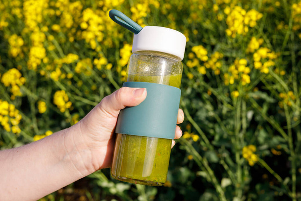  egy sétáló nő kezében egy üveg elkészített gyümölcs és zöldség. smoothie, finom zöld ital. Virágzó sárga virágok a háttérben. Koncepció, egészséges életmód, táplálkozás  - Fotó, kép