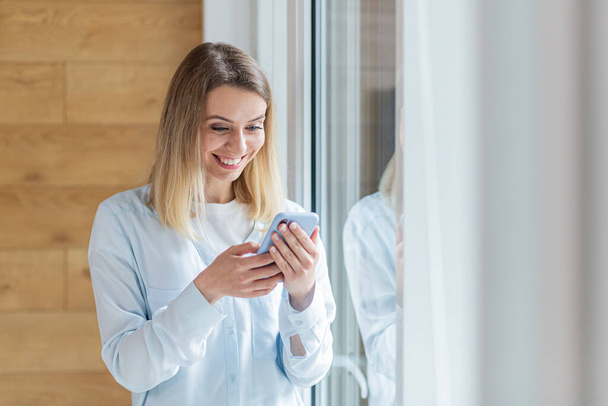 young happy woman saw the good news on smartphone, standing by the window in office or at home indoors. Female with delight holds a mobile phone in his hands reads the news of the day, victory or win - Φωτογραφία, εικόνα