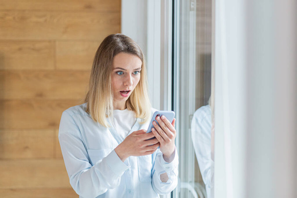 young woman saw bad news on smartphone, standing by the window in the office or at home indoors. The female anxiously holds a mobile phone in her hands, reads the news of the day - Φωτογραφία, εικόνα