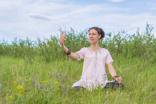 Молода жінка сидить на зеленому газоні в положенні лотоса і фотографує себе на смартфоні
. - Фото, зображення