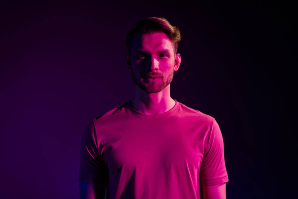 Portrait of attractive serious content guy wearing t-shirt isolated over dark neon light violet color background - Photo, Image