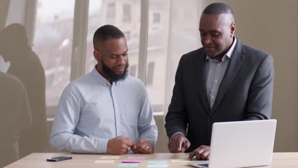 Two african american business men afro mature boss leader and black ethnic manager worker working together standing at table with laptop in office brainstorming discussing ideas write on sticky notes - Footage, Video