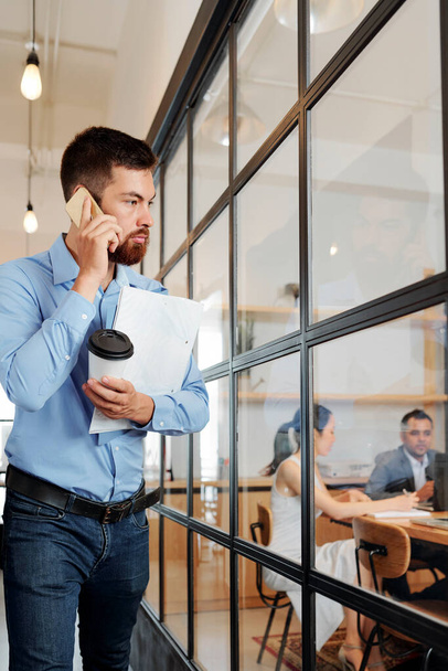 Серйозний молодий бородатий підприємець стоїть за межами кімнати переговорів зі скляними стінами і розмовляє по телефону з колегами
 - Фото, зображення