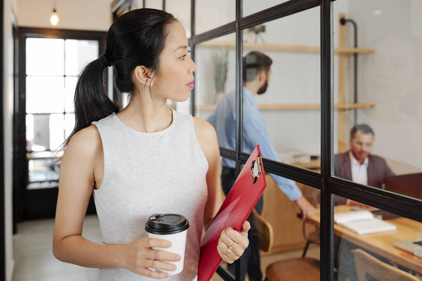 Hübsche junge Geschäftsfrau mit Tasse Kaffee und Aktenmappe kommt am Besprechungsraum mit Glaswänden vorbei und schaut sprechende Mitarbeiter an - Foto, Bild