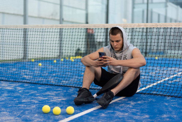 Mladý muž s městským stylem si dává pauzu s telefonem vlaky po hraní pádlo tenis na venkovním hřišti - Fotografie, Obrázek
