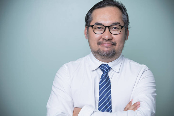 自信の高いアジア系のビジネスマンは、白いシャツを着て、青いネクタイをして、カメラの笑顔を見て眼鏡をかけて、腕を緑色の背景に反対側に立っています。自信のある男. - 写真・画像