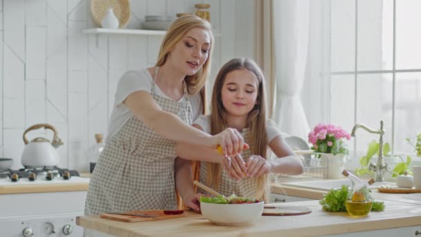 Aikuinen valkoihoinen nainen äiti ja tyttö lapsi tytär käyttävät esiliinoja seistä kotona keittiössä pöydän lähellä valmistella salaattia purista keltaisia sitruunoita lautasella kaatamalla vihanneksia ainesosia, perheen ruoanlaitto - Materiaali, video