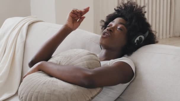 Calm happy young black race woman in headphones sitting on comfortable sofa with eyes closed listening favorite music modern rhythmic pleasant composition enjoy peaceful mood wearing earphones at home - Video, Çekim