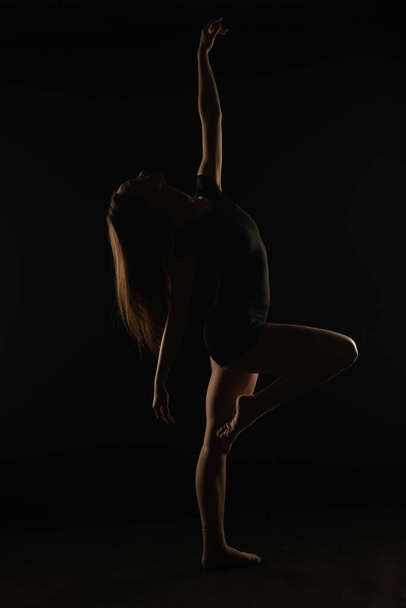 Young ballerina in studio is bending her back while standing on one leg, silhouette - Fotoğraf, Görsel