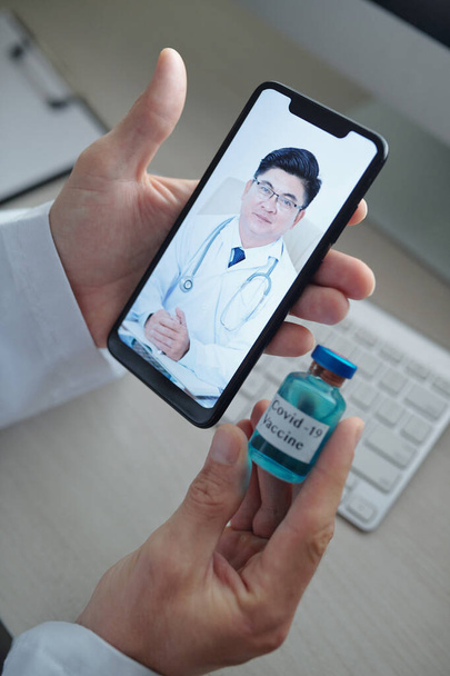 Manos del médico sosteniendo un vial de vacuna contra el coronavirus cuando llama por video a su colega de otro hospital - Foto, imagen