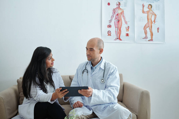 praktičtí lékaři sedící na pohovce v ordinaci lékařů a diskutující seznam symptomů pacientů na tabletu - Fotografie, Obrázek