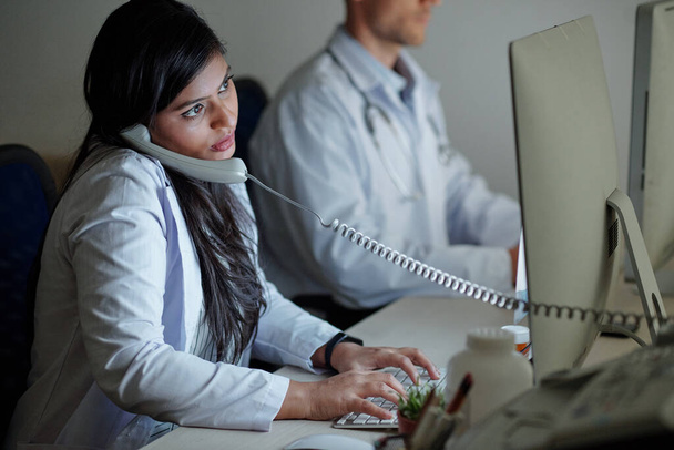 Ciddi genç Hintli kadın doktor hastasıyla telefonda konuşuyor ve şikayetlerini ve belirtilerini bilgisayar ekranına kaydediyor. - Fotoğraf, Görsel