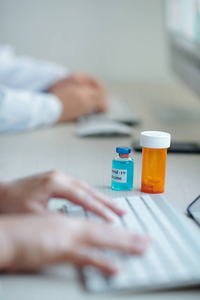 Вакцина проти коронавірусу та пляшка таблеток на столі перед лікарем, який працює на ноутбуці
 - Фото, зображення