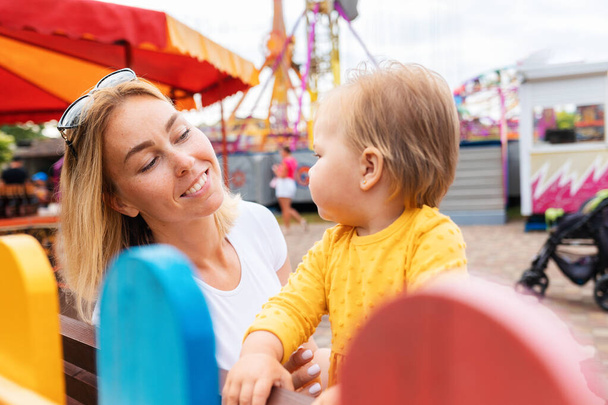 Portrait of smiling caucasian young mother with a little daughter. In the background an amusement park. Concept of summer holiday. - Fotografie, Obrázek