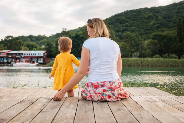 桟橋に座っている母親と赤ん坊の女の子。バックビュー。家族の夏休みの概念. - 写真・画像