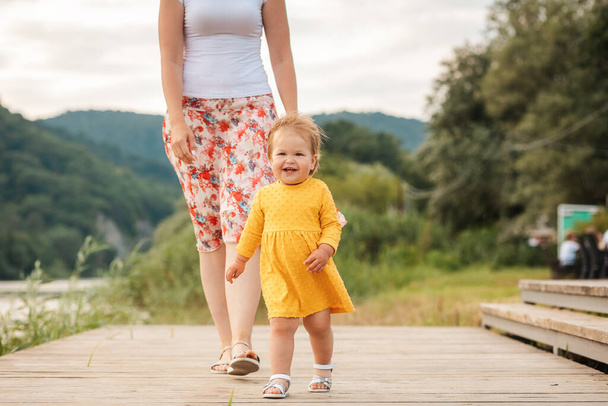 Παιδική ηλικία. Ευτυχισμένο καυκάσιο κοριτσάκι με κίτρινο φόρεμα που περπατάει στην προβλήτα του ποταμού με τη μητέρα της. Εξωτερικά. Έννοια της ημέρας των παιδιών. - Φωτογραφία, εικόνα