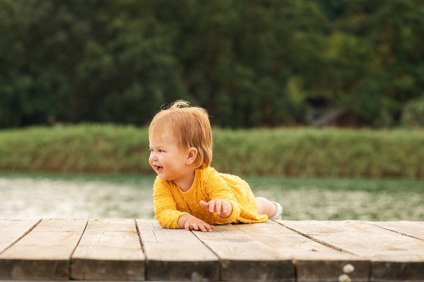 Summertime. Cute happy baby girl lie on the pier by the river. Outdoors. The concept of a happy childhood. - Photo, Image