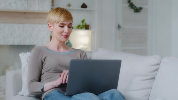 Αρκετά χαρούμενη νεαρή γυναίκα ξαπλωμένη σε άνετο καναπέ με φορητό υπολογιστή σε γύρους, εργάζεται μακριά από το σπίτι, γράφοντας e-mail, απολαμβάνοντας online ψώνια ή επικοινωνώντας στα κοινωνικά δίκτυα, αίσθημα χαλάρωσης το Σαββατοκύριακο - Πλάνα, βίντεο