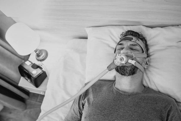 Monochromatyczny portret śpiącego mężczyzny z chronicznymi problemami z oddychaniem rozważa użycie maszyny CPAP w łóżku. Opieka zdrowotna, Obturacyjna terapia bezdechu sennego, CPAP, koncepcja chrapania - Zdjęcie, obraz