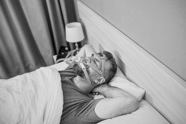 Monochromatický portrét Spícího muže s chronickými dýchacími potížemi zvažuje použití CPAP stroje v posteli. Zdravotní péče, obstrukční spánkové apnoe terapie, CPAP, chrápání koncept - Fotografie, Obrázek