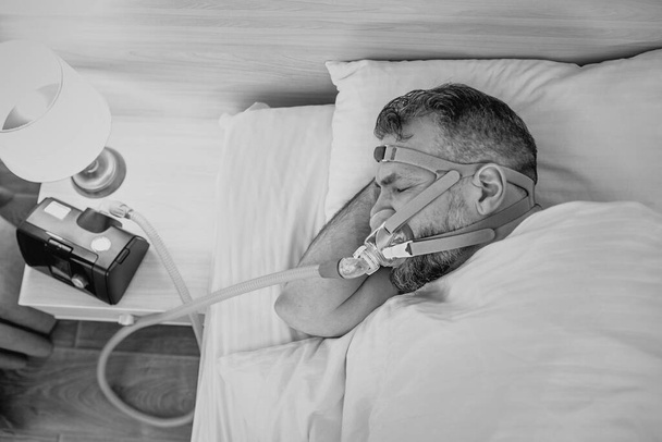Monochrome portrait of Sleeping man with chronic breathing issues considers using CPAP machine in bed. Healthcare, Obstructive sleep apnea therapy, CPAP, snoring concept - Fotó, kép