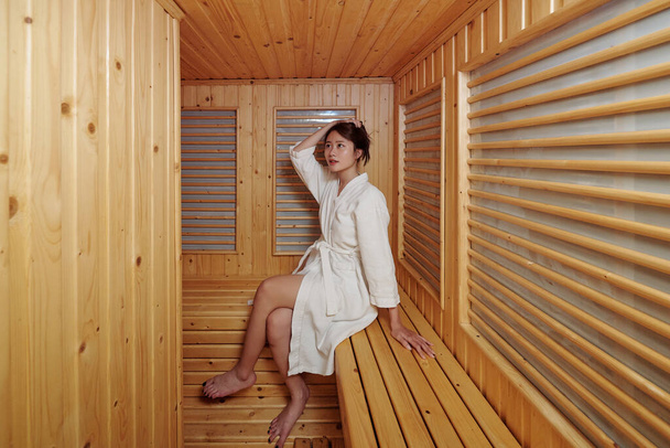 Όμορφη νεαρή γυναίκα σε μπουρνούζι χαλαρώνοντας σε ζεστό σάουνα του ξενοδοχείου spa - Φωτογραφία, εικόνα
