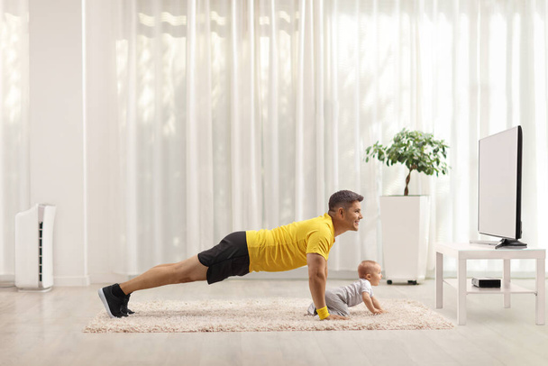 Padre ejercitándose frente a la televisión y el bebé arrastrándose sobre una alfombra aislada sobre un fondo blanco - Foto, Imagen