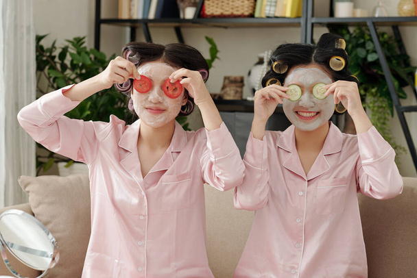 Cheerful young women with moisturizing masks on faces covering eyes with tomato and cucumber slices - Photo, Image