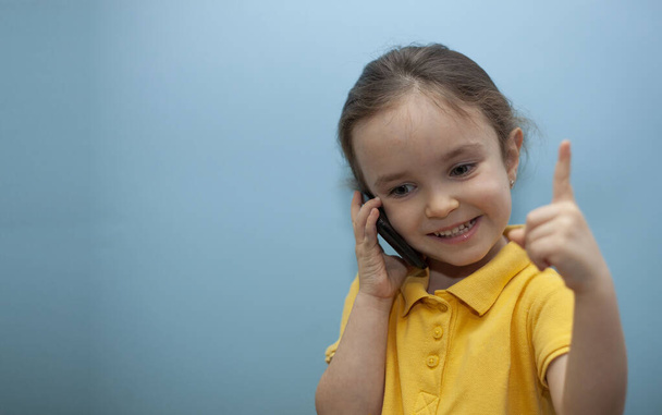 Dziewczynka w żółtej koszulce na niebieskim tle. Dziecko rozmawia przez telefon i patrzy w kamerę. - Zdjęcie, obraz