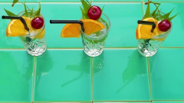 Virkistävä kesän juomia kristalli lasit lehdet kirsikka oranssi vihreä laatta tausta. Makea kylmä luonnollinen cocktail kuuma kesäpäivä. Limsa vesi hedelmät marjat minttu tonic Party loma ylhäältä näkymä - Materiaali, video