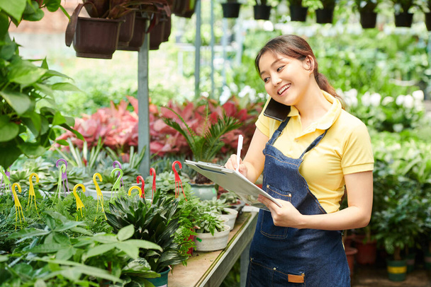 Χαμογελώντας όμορφη κηπουρική εργαζόμενος κέντρο μιλάμε στο τηλέφωνο με τον πελάτη και κρατώντας σημειώσεις στο έγγραφο - Φωτογραφία, εικόνα