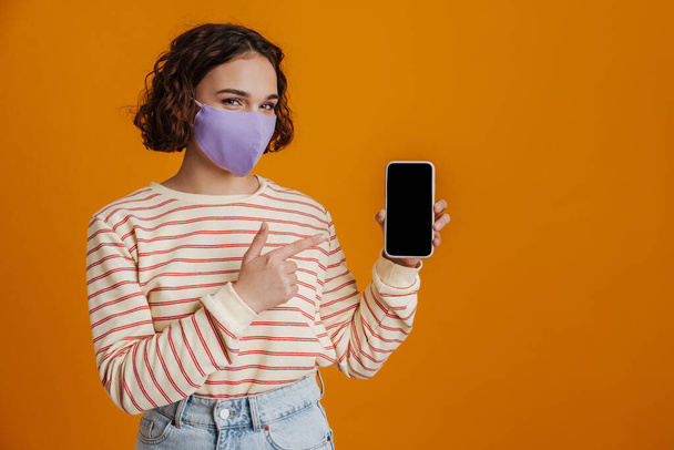 Young confident woman in facial medical mask showing blank screen mobile phone isolated over yellow background - Foto, Imagen