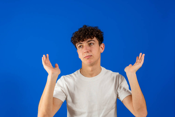 Attraktiver junger Mann mit lockigem Haar und im Zweifel die Hände in die Höhe gereckt, isoliert auf blauem Studiohintergrund. Erstaunliches Konzept - Foto, Bild