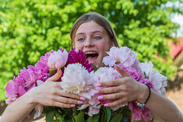 Beautiful and happy young girl in the summer with a beautiful bouquet of peonies in nature. Close up portrait outdoors - Foto, Imagem