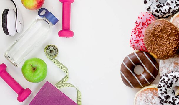 Choose between a healthy lifestyle and junk food. Dumbbells, tape measure, apple, bottle and donuts on a white background. - Photo, image
