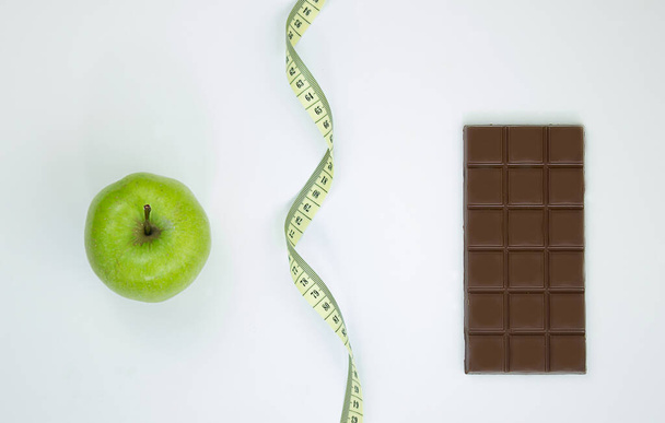 выбирать между здоровым образом жизни и нездоровой пищей. лента яблочного сантиметра и шоколад на белом фоне - Фото, изображение