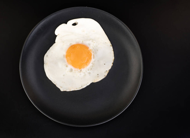 Fried egg lies on a black plate on a black background isolated - Fotografie, Obrázek
