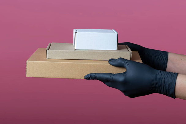 Concept hands in medical black gloves hold cardboard boxes of different sizes, isolated on a pink background. Delivery, moving, packaging and gifts. layout - Фото, зображення