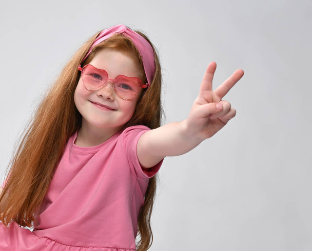 Excited red-haired girl in stylish sunglasses and summer fashion outfit showing victoria sign with her fingers while shooting a studio portrait to the camera - Foto, Bild