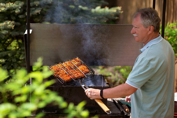 Senior man grilling in the backyard of his country house in sunny summer day. - Photo, Image