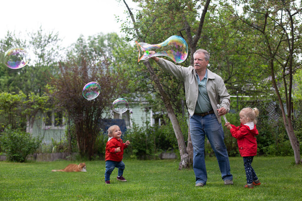 Idősebb férfi játszik az unokáival a kertben. Nagyapa szappanbuborékokat csinál. - Fotó, kép