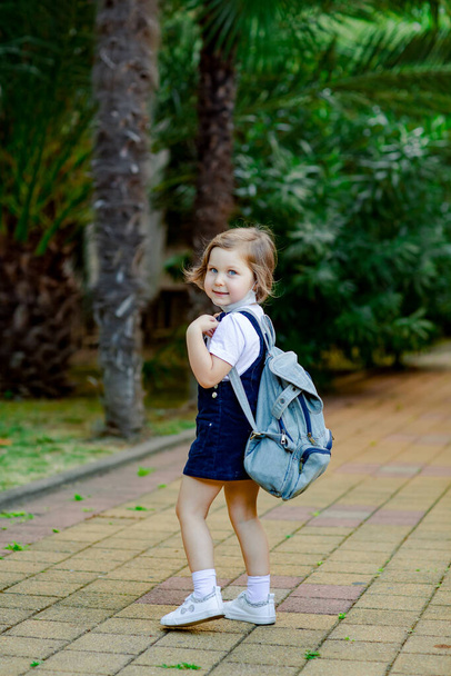 a little girl, a schoolgirl, goes to school in the park with a denim backpack, and in a medical mask - Fotoğraf, Görsel