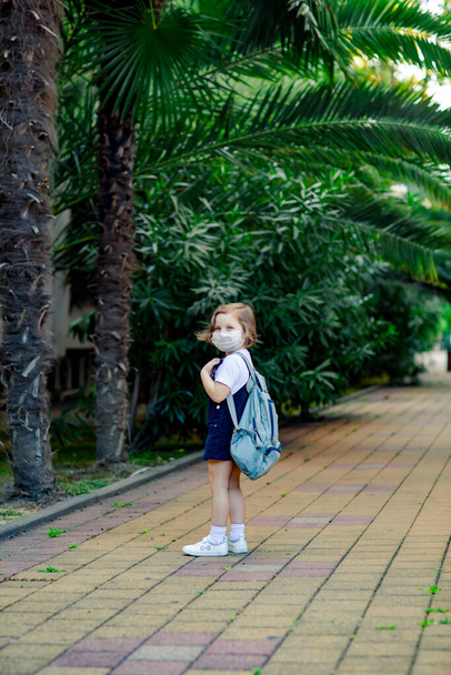 a little girl, a schoolgirl, goes to school in the park with a denim backpack, and in a medical mask - Foto, afbeelding