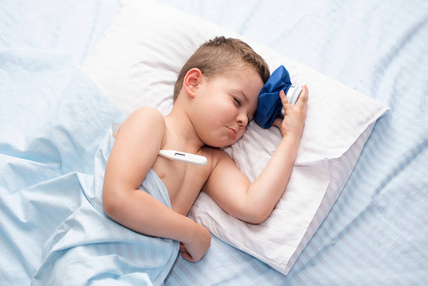 Nemocný malý chlapec s bolestí hlavy leží v posteli a měří teplotu a chladí hlavu studenou krabičkou - Fotografie, Obrázek