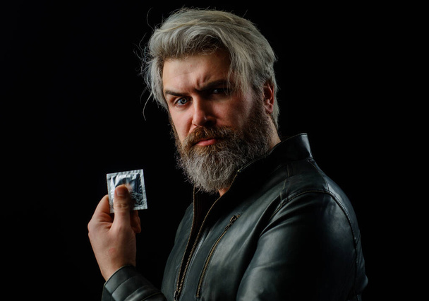 Contraception and healthy lifestyle concept. Bearded Man in leather jacket with condom. Preventive protection for sex. Sex education. - Photo, Image