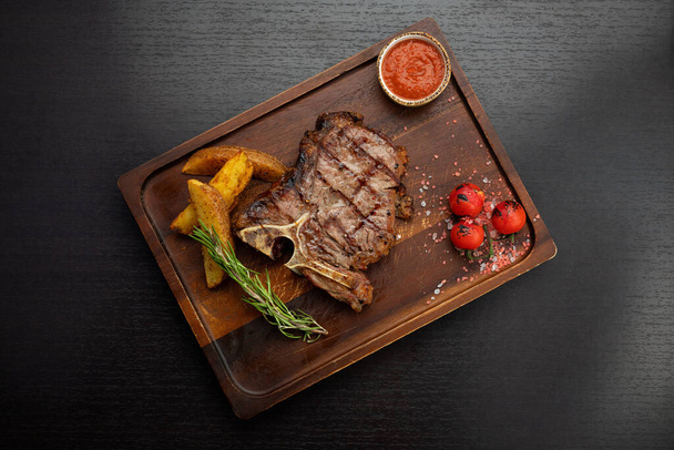 T-bone steak op een houten bord met aardappelen, saus, kersenplukkers, op een donkere tafel - Foto, afbeelding