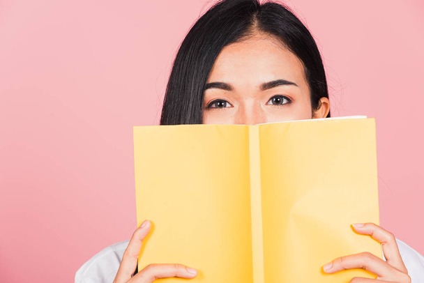 Portrait of beautiful Asian young woman teen smile covering her face with yellow book, female person hiding behind an open book show only eyes, studio shot isolated on pink background - Foto, imagen